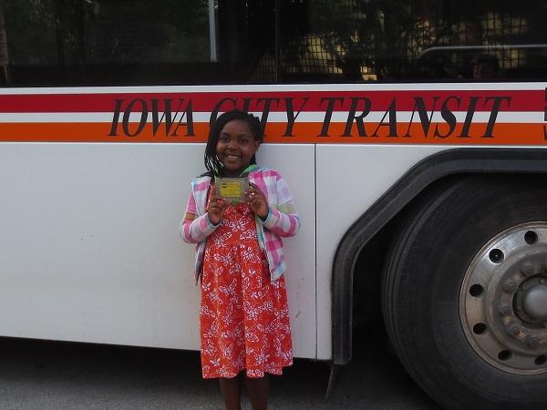a young child with brown skin and a pink dress holds a library card while standing in front of a city bus