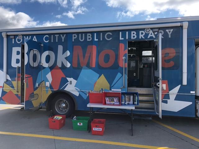 ICPL Bookmobile parked in front of a blue sky, with doors open and bins of books set out for browsing. 